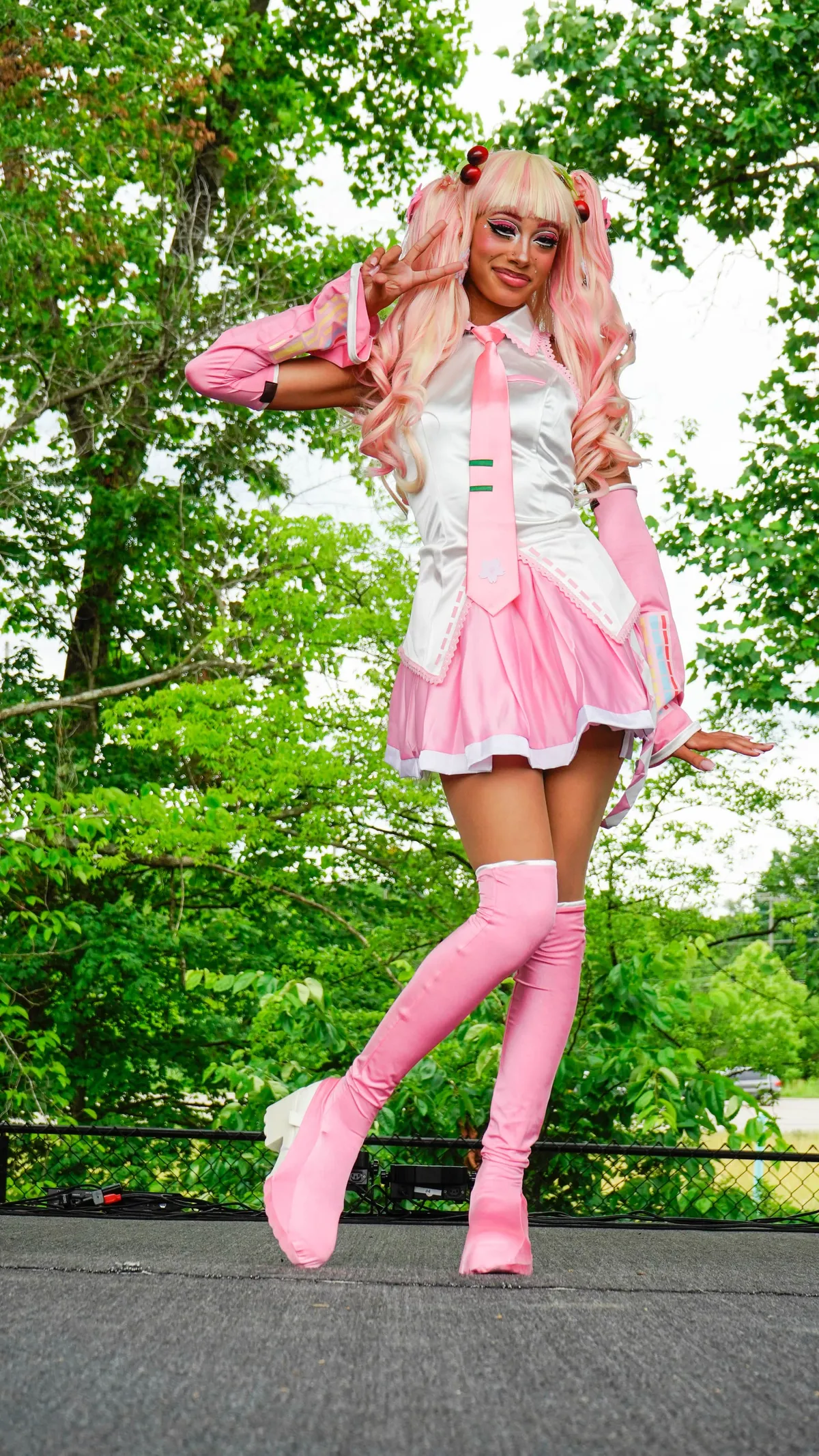 A cosplayer in a pink-and-white idol costume with long pink twin-tail curls, arm warmers and pink thigh-high boots poses with a peace sign on an outdoor stage before a leafy tree line at Knox Pop Con.